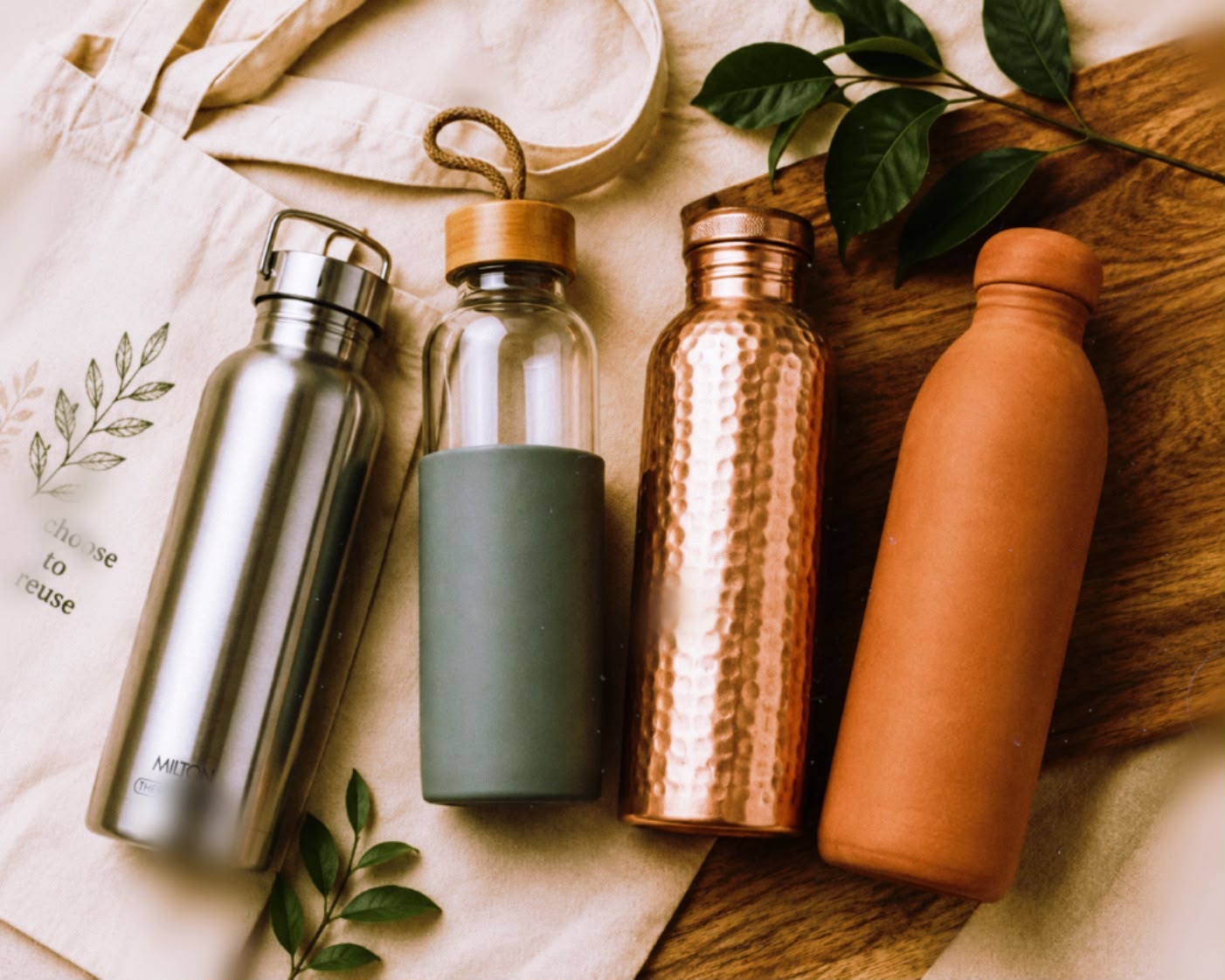 Reusable water bottles made of stainless steel, glass, copper, and terracotta on a neutral beige background with green leaves and a cotton tote bag.